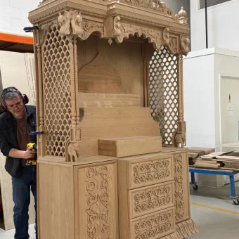 A large, floor-to-standing custom wooden Pooja Ghar (mandir) featuring intricate lattice-work screens, a tiered decorative roof, and hand-carved floral patterns on the base cabinets.