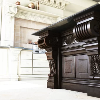 A heavy, dark-stained kitchen island featuring architectural island posts with deep scroll carvings and intricate classical moldings, contrasting against a bright white marble floor.