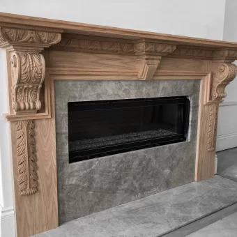 A light wood fireplace surround in a living room, featuring hand-carved decorative corbels, floral appliqués, and a classic fluted design surrounding a modern gray stone hearth.