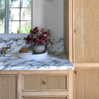 Close-up of custom kitchen cabinetry featuring vertical reeded (tambour) door panels in a natural light oak finish, paired with a luxurious marble countertop and backsplash for a modern organic look.