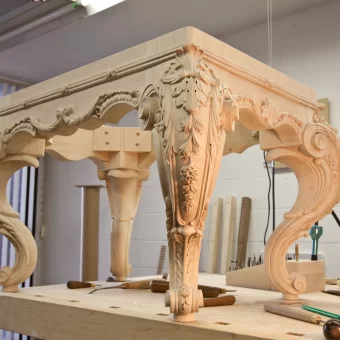 A custom-made wooden table in progress, featuring cabriole legs and a thick apron entirely covered in deep-relief carvings of flowing flowers and leaves.