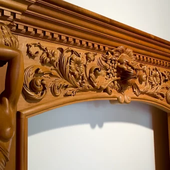 A highly detailed wooden fireplace mantel featuring a central lion head carving surrounded by floral scrolls and flanking female figures in a Baroque architectural style.