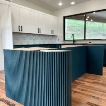 A modern kitchen featuring a curved island base wrapped in flexible, vertical-slat maple tambour panels, ready for custom paint or stain.