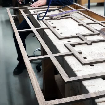 A large-scale architectural wood frame featuring an intricate geometric mullion design, precision-crafted from a single piece of solid wood for a seamless finish on a workshop table.