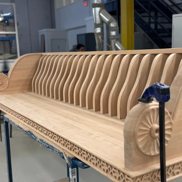 A long, custom-built wooden bench in a workshop setting, featuring a modern vertical slat backrest and a highly decorative, traditional hand-carved apron along the base.