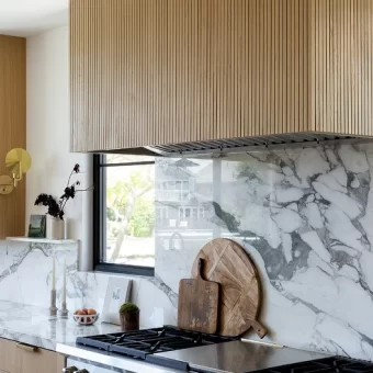 A bright, modern kitchen featuring a custom range hood wrapped in flexible white oak tambour panels. The vertical wood slats add texture and warmth to the minimalist white marble and stainless steel surroundings.